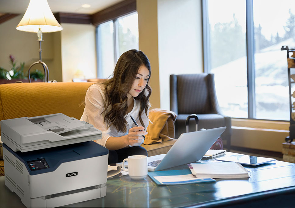 Xerox C235 Multifunction Printer in Home Office Woman working from home using laptop next to Xerox C235 multifunction printer, ideal for hybrid work and home office printing.