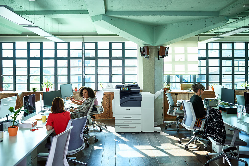 Two women collaborating in an office using a tablet next to a Xerox VersaLink C625 multifunction printer